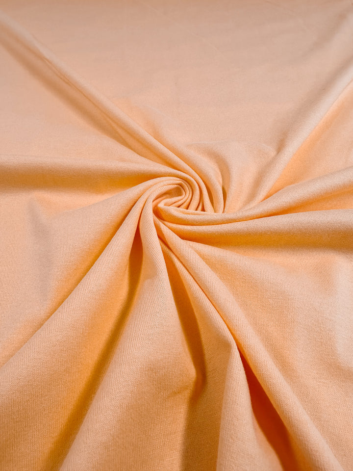 Close-up of Cotton Jersey - Papaya - 160cm by Super Cheap Fabrics with a spiral fold in the center, creating gentle creases and shadows on the smooth surface—a perfect medium weight fabric reminiscent of soft peach-colored textures.