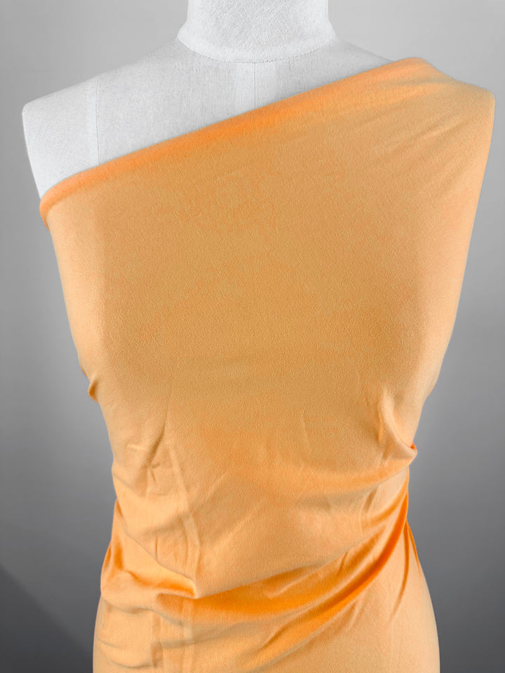 A Super Cheap Fabrics mannequin is adorned with a single piece of Papaya Cotton Jersey, 160cm wide, draping around its shoulder and torso. The smooth texture and subtle sheen resemble papaya's hue, striking against the white mannequin.