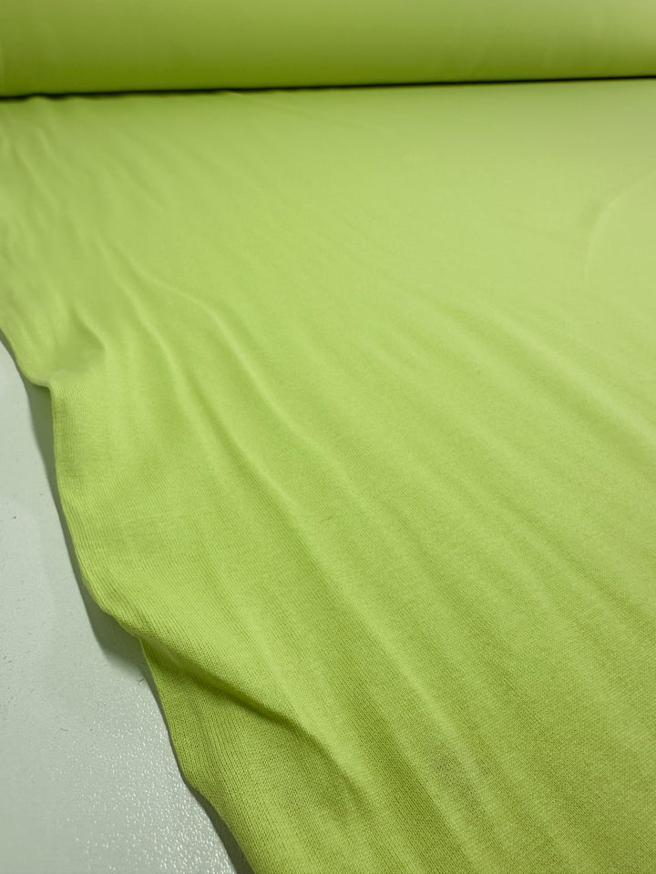 A roll of Super Cheap Fabrics' Cotton Jersey in Daiquiri Green - 170cm is partially unrolled on a flat surface, revealing its soft, smooth texture and uniform color.