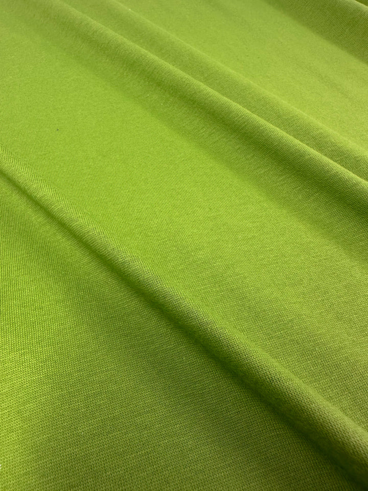 A close-up of the Cotton Jersey - Titanite by Super Cheap Fabrics reveals a green, textured fabric with gentle folds and ridges. The soft material has a smooth surface highlighted by the lighting, showcasing its subtle contours. Width: 160cm.