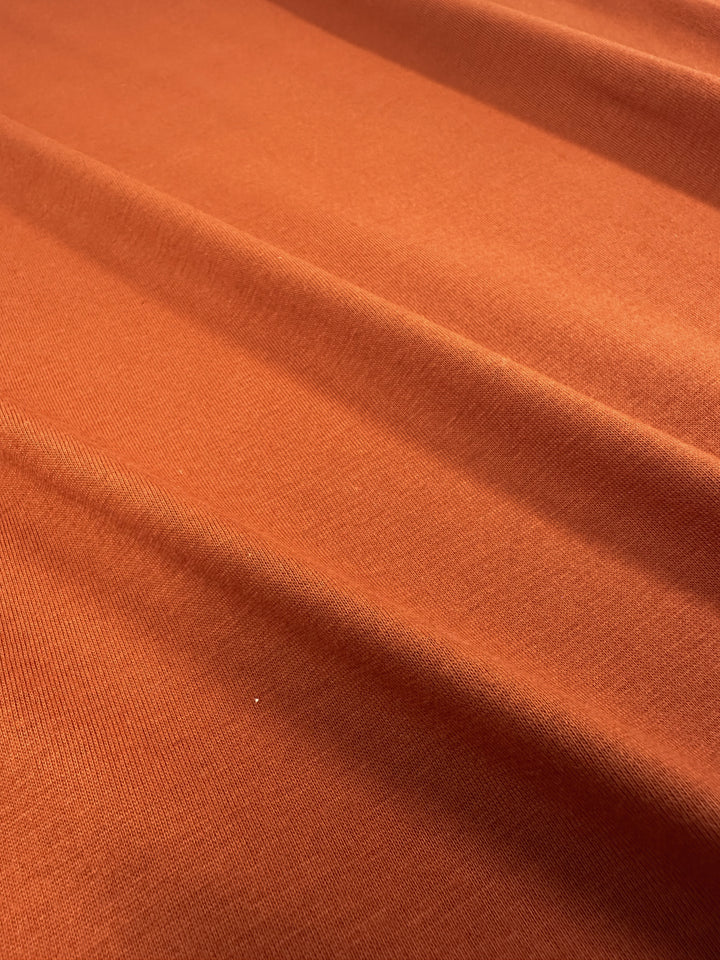 A close-up of the soft Cotton Jersey - Apricot Orange by Super Cheap Fabrics shows gentle folds and a wavy texture. The light highlights its smooth, slightly shiny finish and rich apricot orange hue. .