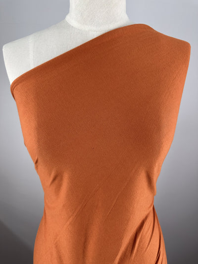 A mannequin showcases Super Cheap Fabrics' Cotton Jersey in Apricot Orange, gracefully styled diagonally over one shoulder against a neutral gray background, highlighting its lightweight texture and elegant drape.