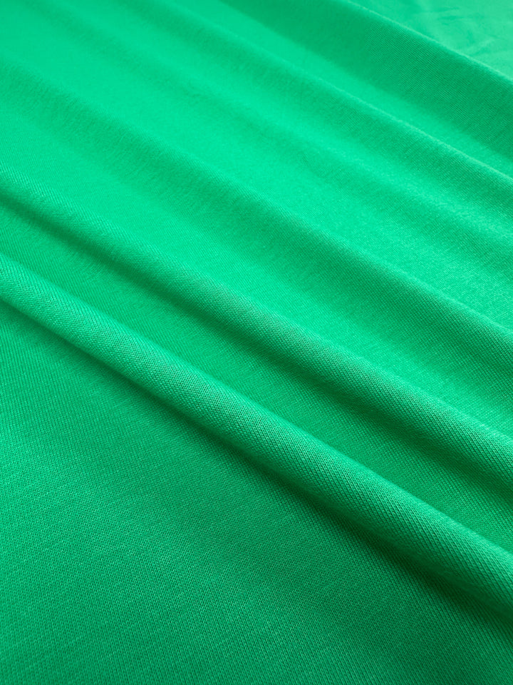 Close-up of Super Cheap Fabrics' Cotton Jersey in Vivid Green (165cm) shows a smooth, soft texture with gentle folds. The slightly stretchy cotton creates subtle shadow and highlight patterns on the surface.