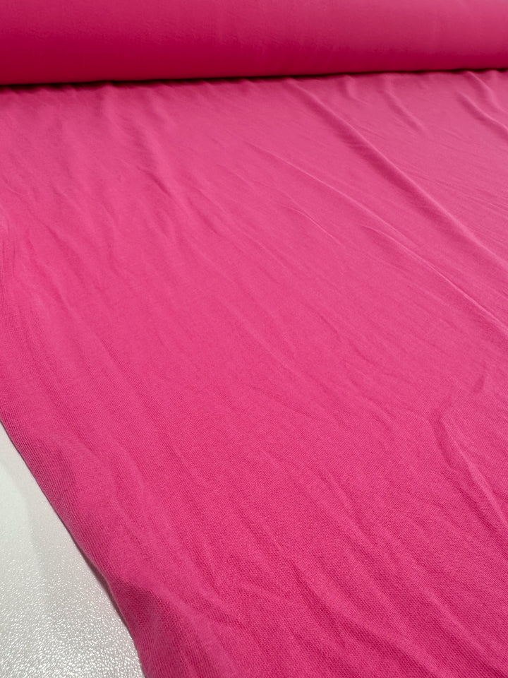 A large roll of Cotton Jersey - Fandango Pink from Super Cheap Fabrics unfurls on a flat surface, displaying its smooth and slightly stretched texture. The vibrant 160cm-wide fabric dominates the image.