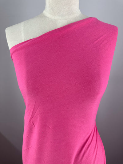 A mannequin elegantly displays Super Cheap Fabrics' Cotton Jersey in vibrant Fandango Pink, featuring an off-shoulder style, set against a gray background.