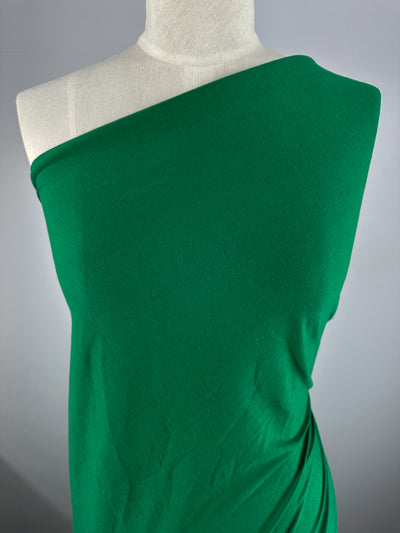 A plain background showcases a mannequin draped in Super Cheap Fabrics' Cotton Jersey in Abundant Green, 165cm, with its smooth 100% cotton texture elegantly covering the torso.