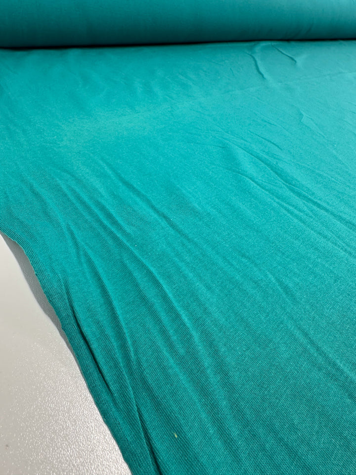 A large roll of turquoise Cotton Jersey - Kayaking by Super Cheap Fabrics is spread out, showcasing its smooth texture with visible wrinkles and folds. This lightweight 155cm fabric is ideal for crafting jumpsuit dresses or upholstery projects.