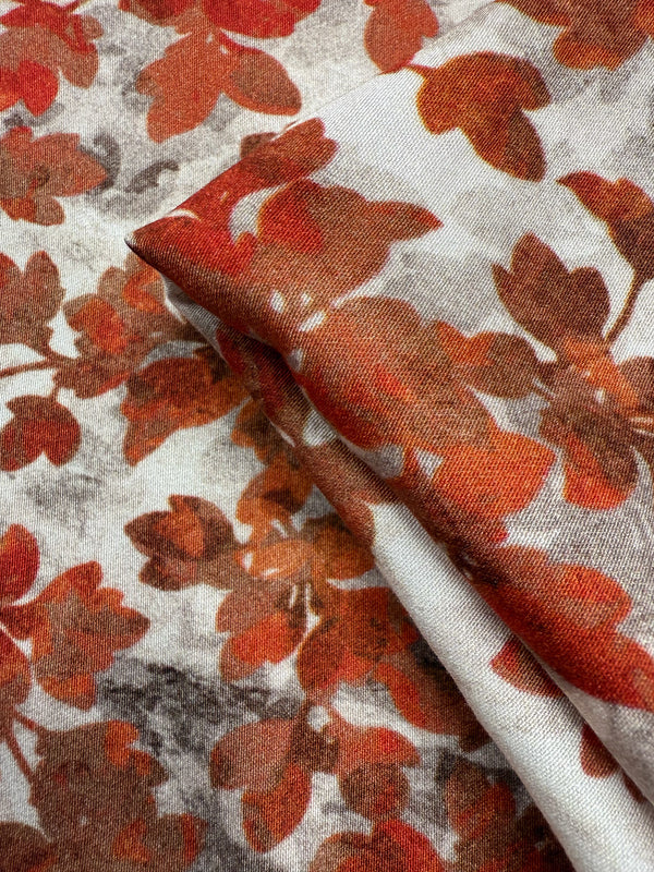 A close-up of Super Cheap Fabrics' Mercerised Rayon - Crimson Canopy (150cm), showing its lightweight, breathable material with a red and orange autumn leaf pattern on a white background, slightly folded.