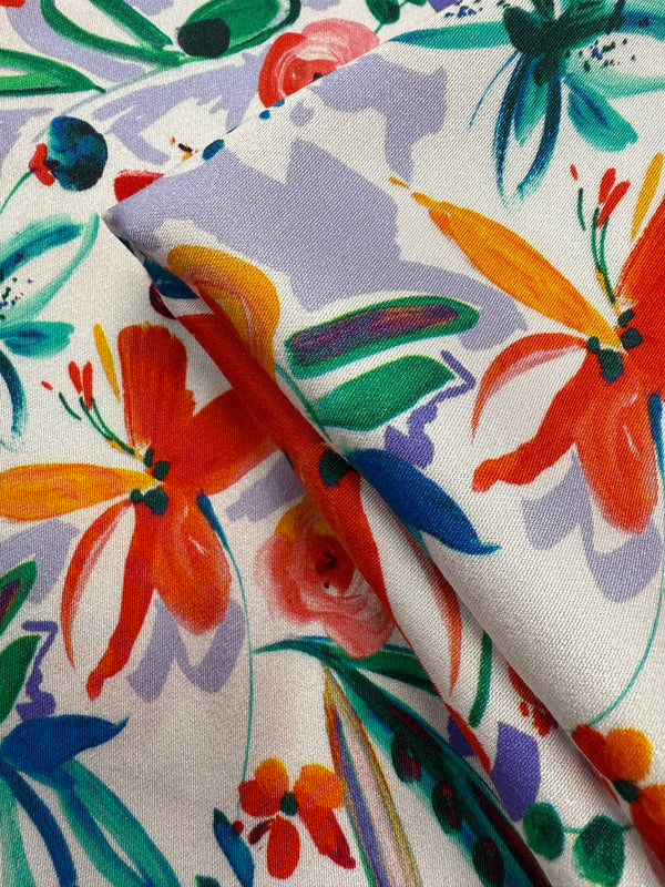 Close-up of Super Cheap Fabrics' Mercerised Rayon - Tropical Brushstroke (150cm) showing bright orange, yellow, green, and pink floral patterns with leaves on a white background. The lightweight fabric is partially folded to reveal its vibrant texture.