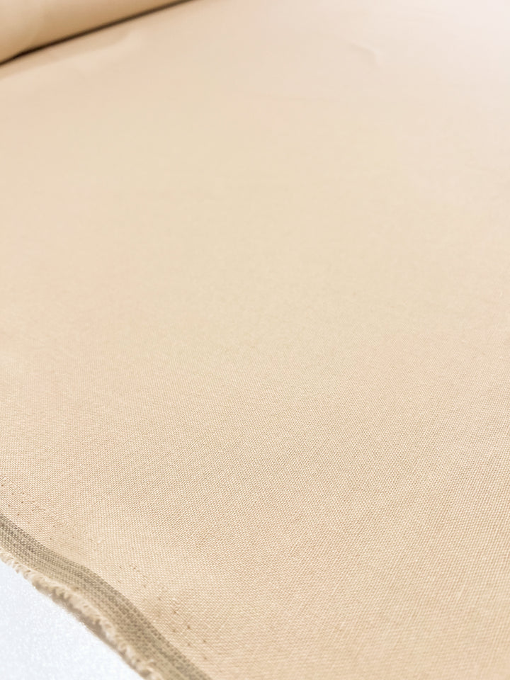 A close-up of the Premium Linen Cotton - Shortbread from Super Cheap Fabrics, featuring a lightweight roll laid out on a flat surface. The material appears smooth and uniform, showcasing a visible selvage edge on one side. The lighting highlights the fabric's fine texture and subtle sheen, making it stand out beautifully in its shortbread color.