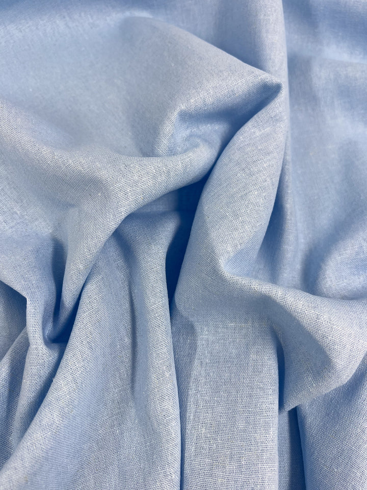 A close-up of Super Cheap Fabrics' Premium Linen Cotton - Clear Sky - 140cm showcases the lightweight and slightly textured linen material. The soft, light blue fabric is elegantly draped, forming gentle folds and shadows that evoke the tranquil beauty of a clear sky, highlighting its fluidity and softness.