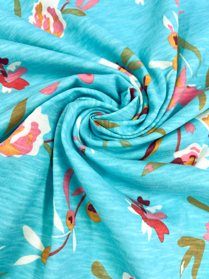 Introducing the Evelyn Printed Jersey from Super Cheap Fabrics – a 170cm wide medium-weight fabric. This material features a beautiful swirl of blue enhanced by a floral pattern in vibrant red, pink, and off-white tones, accented with green leaves. Its durable stretch ensures a spiral effect that highlights its vivid colors and intricate design.