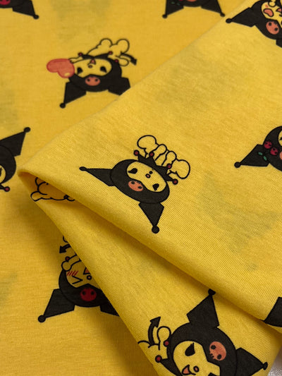 The Super Cheap Fabrics "Printed Jersey - Pixy - 145cm" features a whimsical design of cartoon characters dressed in black jester-like outfits, adorned with yellow horned hats and rosy pink cheeks. One lively character carries a lollipop, and the fabric is folded to highlight the playful pattern from different perspectives.