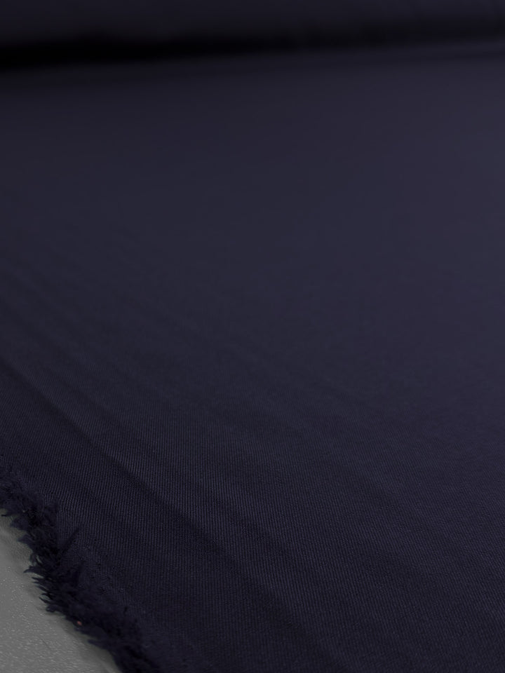 A close-up of Super Cheap Fabrics' Liquid Twill - Black Iris (150cm), laid flat to show its dark navy hue, subtle texture, and frayed edge in the foreground.