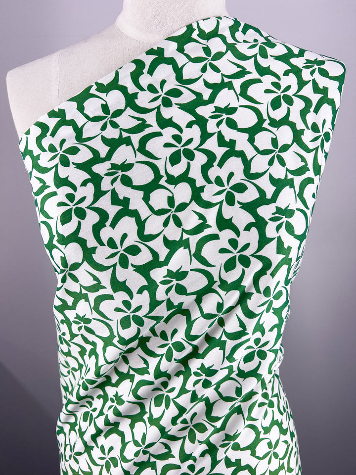 A mannequin showcases Super Cheap Fabrics' Designer Rayon - Green Bee (140cm) with a green and white floral pattern of stylized flowers and leaves, vibrantly standing out against a neutral gray background.