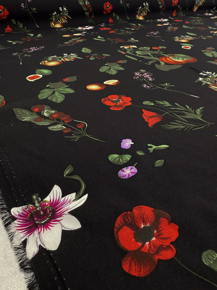 A close-up of Super Cheap Fabrics' Designer Rayon - Dark Orchard Bloom (145cm), showcasing a vibrant floral and fruit print with red poppies, purple orchids, green leaves, and assorted fruits on a black background.