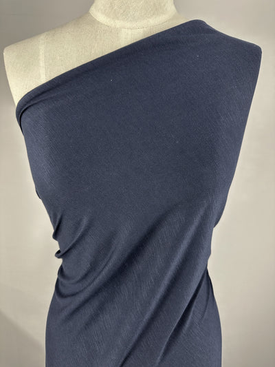 A navy blue, lightweight Bamboo Jersey - Navy - 150cm from Super Cheap Fabrics is draped elegantly over a mannequin with one shoulder exposed, showcasing the texture and drape of the material. The background is plain, emphasizing the focus on this environmentally responsible fabric.