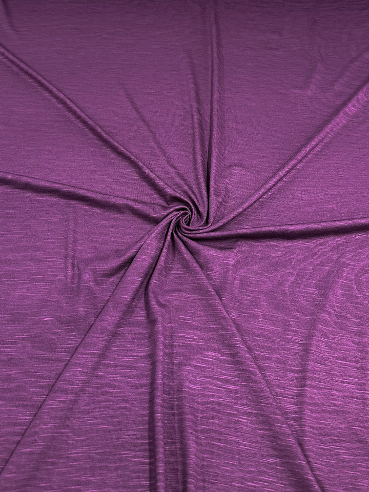 A close-up view of a piece of Bamboo Jersey - Deep Purple - 150cm fabric by Super Cheap Fabrics with a slightly wrinkled texture. The fabric is arranged in a circular, twisted pattern in the center, creating radial folds extending outward. The material appears soft, slightly shiny, and environmentally responsible.