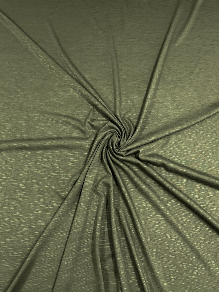 A piece of olive green, eco-friendly Bamboo Jersey - Olive - 150cm from Super Cheap Fabrics with a subtle, slightly shiny texture. The durable fabric is arranged in a swirling pattern, creating folds and creases that radiate outward from the center.