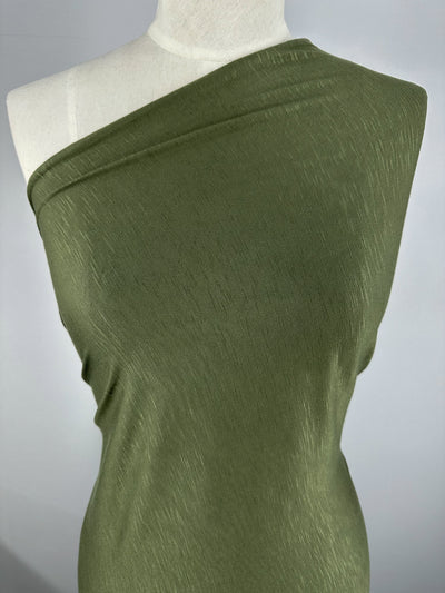 A close-up view of a mannequin dressed in a green, one-shoulder garment. The Bamboo Jersey - Olive - 150cm from Super Cheap Fabrics appears to be smooth with a slight sheen. The background is a plain, neutral color, emphasizing the draped design and eco-friendly nature of the durable green clothing.