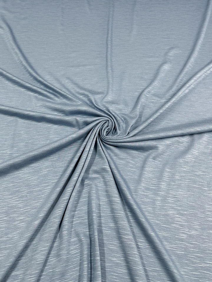 A close-up of a piece of light grey bamboo fabric with a swirl pattern at its center. The material appears soft and lightweight with subtle texture, fine lines, and slight sheen. Environmentally responsible and stylish, the Bamboo Jersey - Powder Blue - 150cm from Super Cheap Fabrics is gently gathered and spiraled, creating radiating folds.
