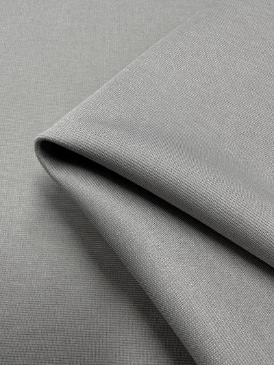 A close-up view of folded gray fabric with a fine, grid-like texture. The material appears smooth and tightly woven, suitable for clothing or upholstery. Crafted from 100% polyester, the medium to heavy weight fabric emphasizes its pliability and durability through the folds. This is the Ponte - Atmosphere - 155cm by Super Cheap Fabrics.
