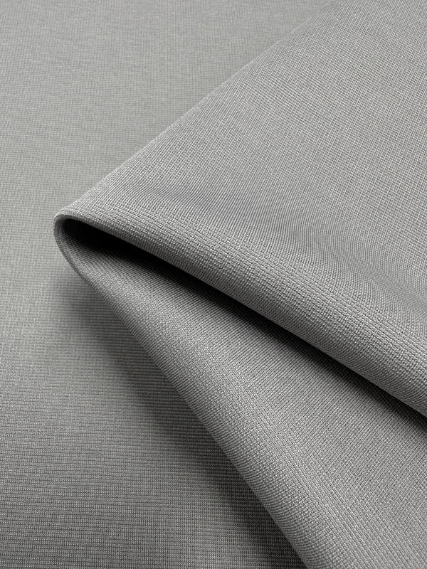 A close-up view of folded gray fabric with a fine, grid-like texture. The material appears smooth and tightly woven, suitable for clothing or upholstery. Crafted from 100% polyester, the medium to heavy weight fabric emphasizes its pliability and durability through the folds. This is the Ponte - Atmosphere - 155cm by Super Cheap Fabrics.