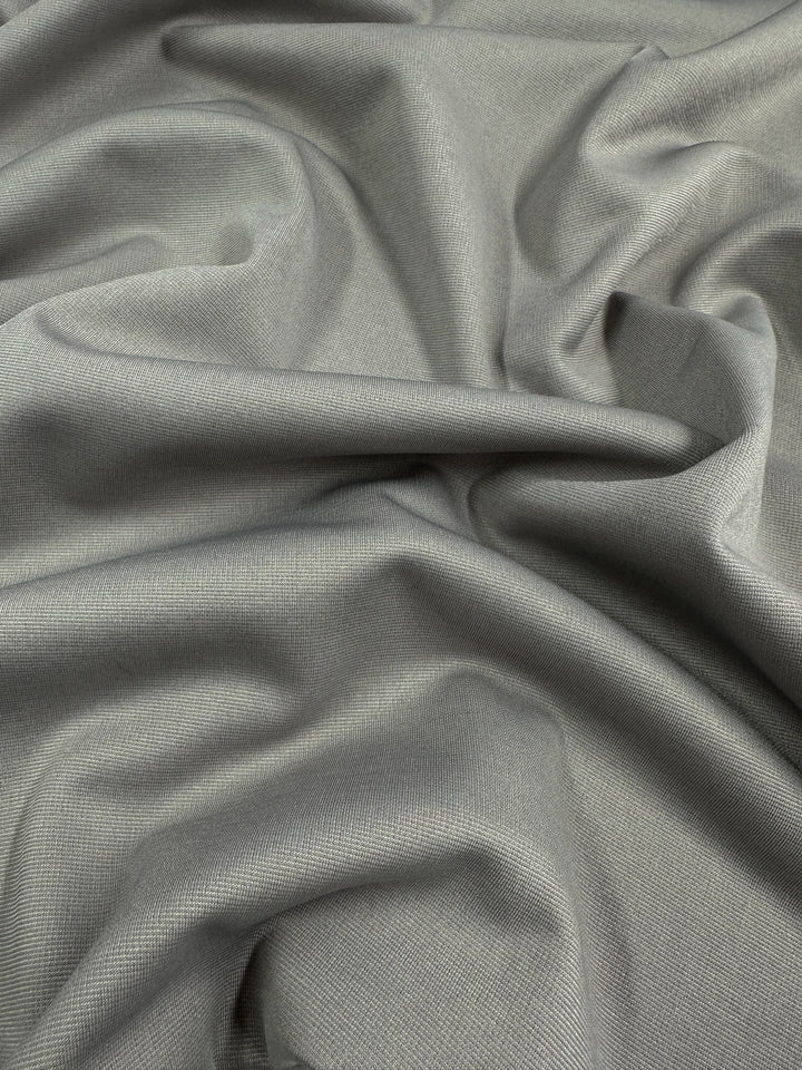 A close-up of a grey, textured fabric with gentle folds and creases. The 100% polyester Ponte - Atmosphere - 155cm from Super Cheap Fabrics has a soft, smooth appearance, light shading that enhances the ripples and contours throughout the surface, and stretches 30%-40%, making it ideal as a medium to heavy weight fabric.