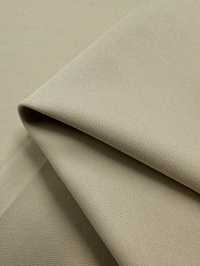 A close-up image of beige Ponte - Fog - 155cm fabric by Super Cheap Fabrics, neatly folded. The material appears to be of medium thickness with a fine, uniform texture. The fabric is shown in smooth, even layers, suggesting it is suitable for various sewing or upholstery projects.