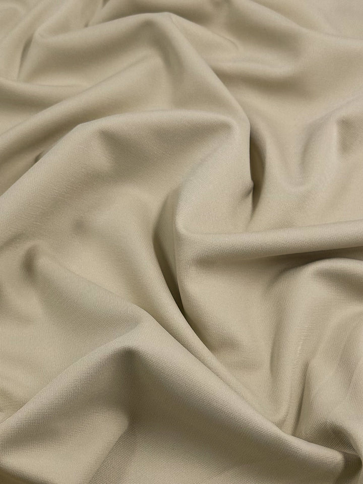 A close-up view of beige, smooth polyester fabric with soft, flowing folds and wrinkles. The texture appears soft and slightly glossy, suggesting a lightweight material often used for medium to heavy weight applications like stretchy pants. This is the Ponte - Fog - 155cm from Super Cheap Fabrics.