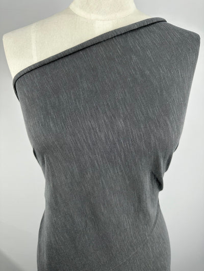 A close-up image of a gray, single-shoulder dress displayed on a mannequin. Made from environmentally responsible Bamboo Jersey - Charcoal - 155cm by Super Cheap Fabrics, the dress has a diagonal neckline that exposes one shoulder, with a simple and smooth texture. The background is plain white.