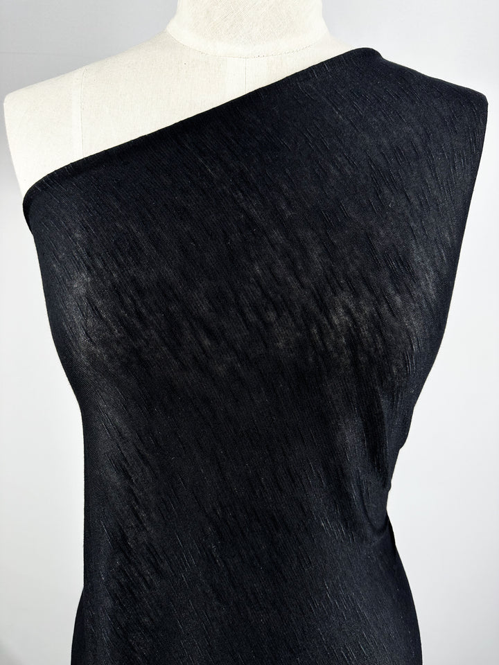 A mannequin draped with black textured Bamboo Jersey - Black - 170cm from Super Cheap Fabrics. The environmentally responsible fabric covers the mannequin's torso diagonally, leaving one shoulder exposed. The background is plain and light grey.