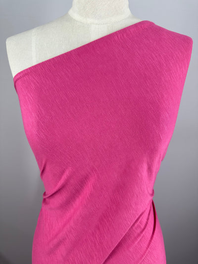 Close-up of a mannequin wearing a vibrant pink, one-shoulder dress made from Super Cheap Fabrics' Bamboo Jersey - Pink - 160cm. The durable fabric is wrapped around the torso, creating a draped effect. The background is a plain gray.