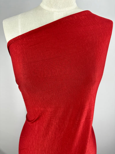 A red, one-shoulder garment draped over a dress form is shown against a plain background. The Bamboo Jersey - Deep Red - 160cm from Super Cheap Fabrics, with its subtle textured pattern, gives it a slightly shiny appearance. The dress form's head and lower portion are not visible.