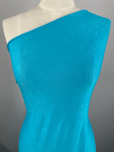 A mannequin is displaying a one-shoulder, fitted turquoise dress against a plain gray background. Made from Super Cheap Fabrics' Bamboo Jersey - Sky Blue - 155cm, the dress has a sleek, textured finish that drapes smoothly over the mannequin's torso while being environmentally responsible.