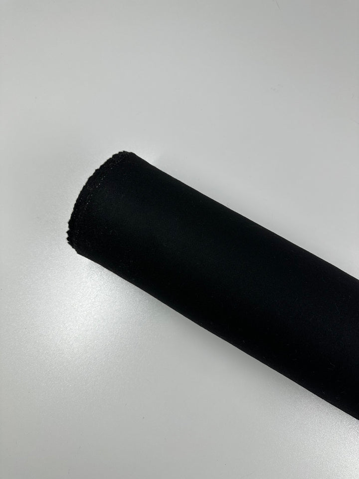 A roll of Virgin Wool - Black - 150cm by Super Cheap Fabrics, made in Italy, is placed diagonally on a white surface. The black material appears thick and smooth, wrapped tightly in a cylindrical shape with one end slightly frayed.