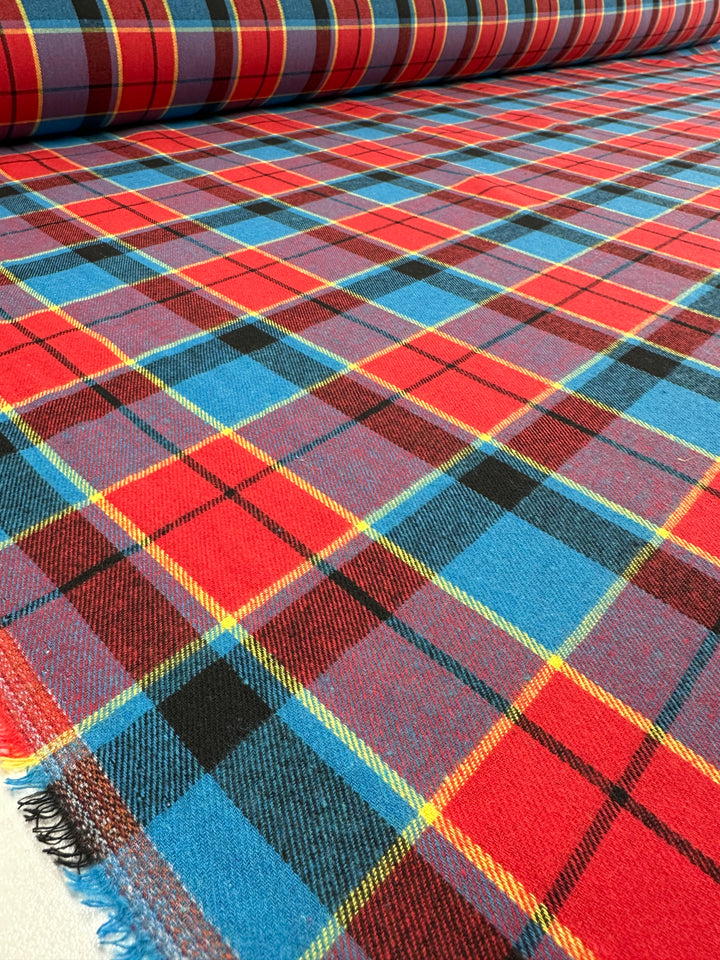 A close-up of a large roll of Super Cheap Fabrics' Suiting - Lolly Clan Watch - 147cm with a vibrant tartan pattern featuring red, blue, yellow, and black stripes. This versatile fabric is laid out flat with a slightly frayed edge visible in the foreground.