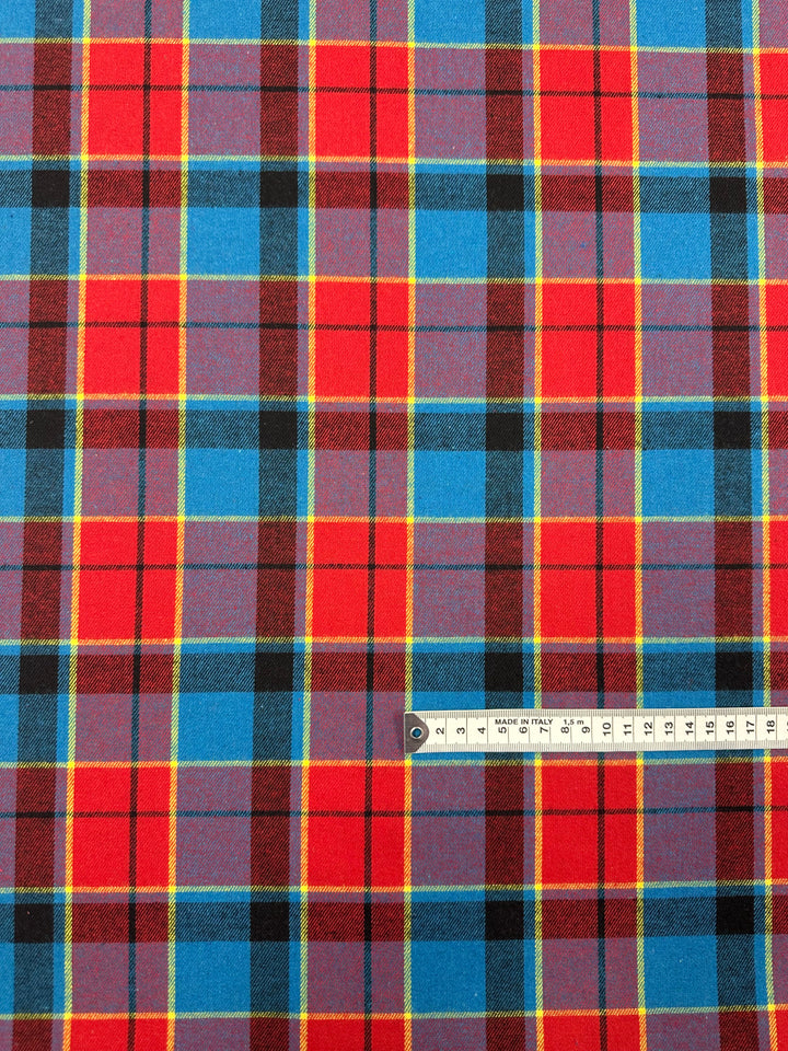 A piece of vibrant tartan twill suiting fabric featuring a pattern of red, blue, yellow, and black intersecting stripes. A measuring tape in centimeters is placed at the bottom, indicating scale. The fabric is called Suiting - Lolly Clan Watch - 147cm by Super Cheap Fabrics.