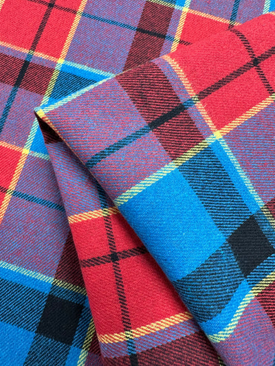 A folded piece of tartan fabric with a pattern of intersecting stripes in red, blue, black, yellow, and green. This versatile fabric displays a traditional plaid design commonly seen in Scottish textiles, making it ideal for formal suits and twill suiting. The product is "Suiting - Lolly Clan Watch - 147cm" by Super Cheap Fabrics.