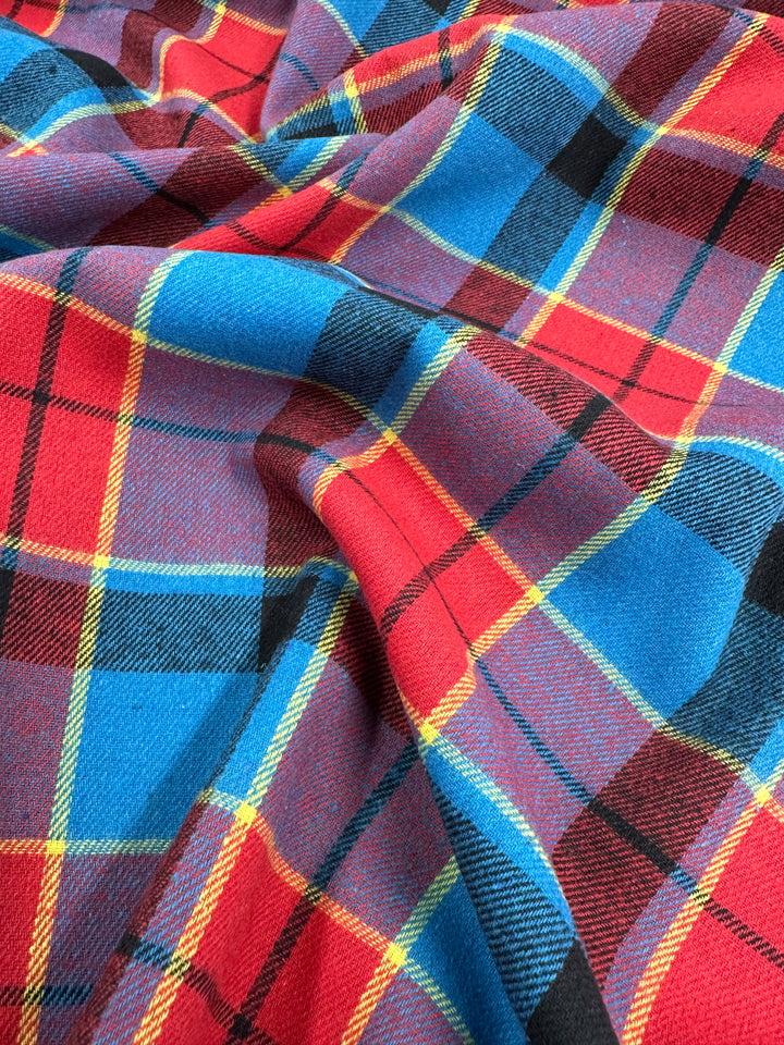 A close-up of a colorful tartan fabric, often found in formal suits, featuring a bold pattern with intersecting stripes of red, blue, black, and yellow. The fabric appears soft and slightly crumpled, showcasing the vibrant and intricate design typical of versatile fabrics like the Suiting - Lolly Clan Watch - 147cm by Super Cheap Fabrics.
