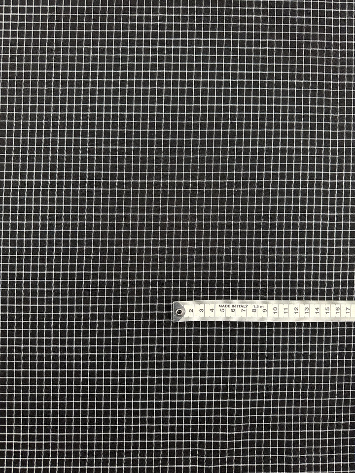 A black fabric with a small white grid pattern, perfect for garment creations or home decor items. A measuring tape is laid horizontally across the bottom right corner, showing measurements in centimeters from 110 to 140. The Linen Rayon Grid - Black - 145cm by Super Cheap Fabrics provides a precise and uniform pattern across the entirety of the fabric.