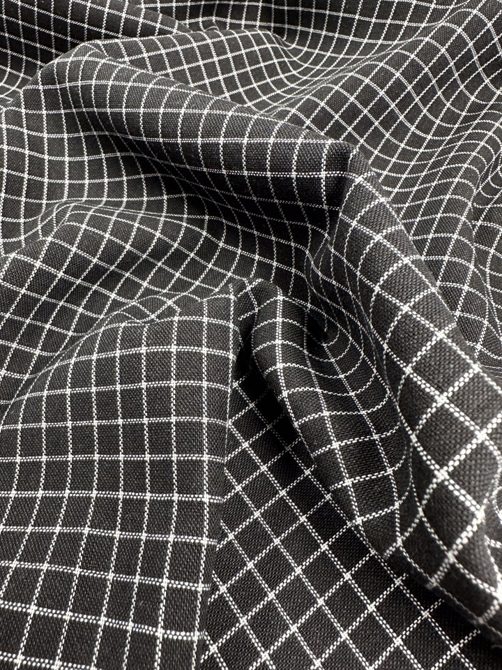 A close-up of Super Cheap Fabrics' Linen Rayon Grid - Black - 145cm with a white grid pattern. The fabric is draped and folded, creating shadows and highlights that emphasize its texture and the geometric lines of the grid, perfect for innovative garment creations or stylish home decor items.