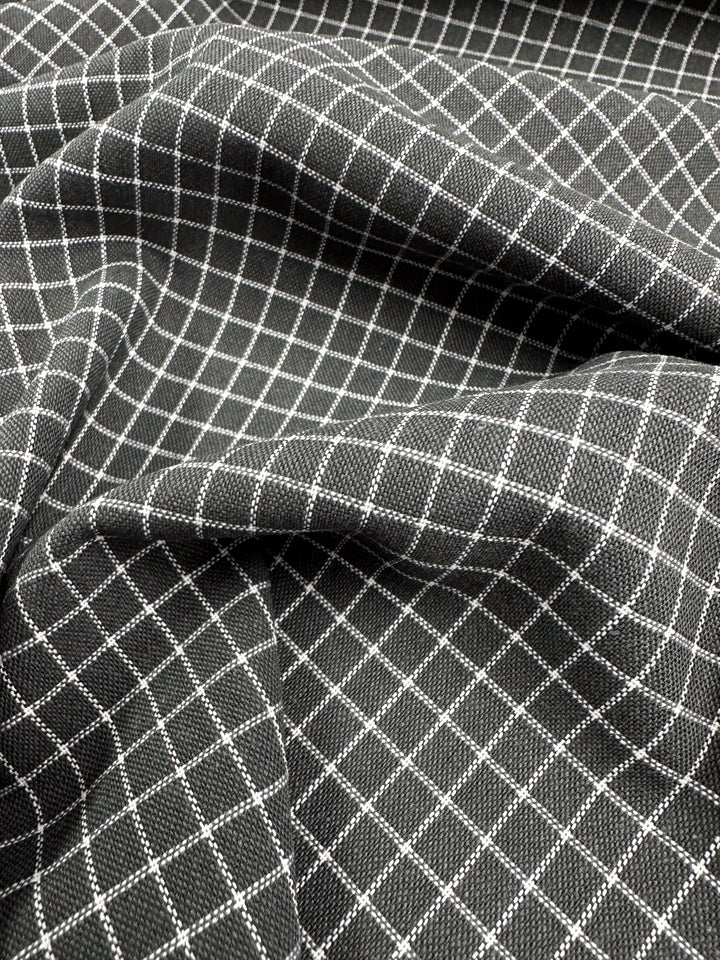 A Linen Rayon Grid - Charcoal - 145cm from Super Cheap Fabrics is draped in soft folds, featuring a dark base color with a white grid pattern of evenly spaced lines forming squares. The material appears textured and has a smooth, slightly reflective surface, perfect for home decor items or garment creations.