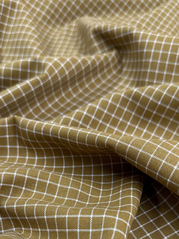 A close-up of a piece of Super Cheap Fabrics' Linen Rayon Grid - Mustard - 145cm featuring a white and tan checkered pattern. The fabric is slightly wrinkled, creating subtle folds and shadows that add depth to the texture, making it perfect for home decor accents.