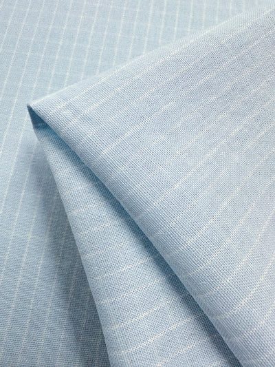A close-up of light blue fabric with a fine white pinstripe pattern. The high-quality Linen Rayon Grid - Baby Blue - 145cm from Super Cheap Fabrics is neatly folded, showcasing both the front and back sides, highlighting its texture and pattern. Ideal for home decor items or garment creations, the pinstripes run parallel across the fabric surface.