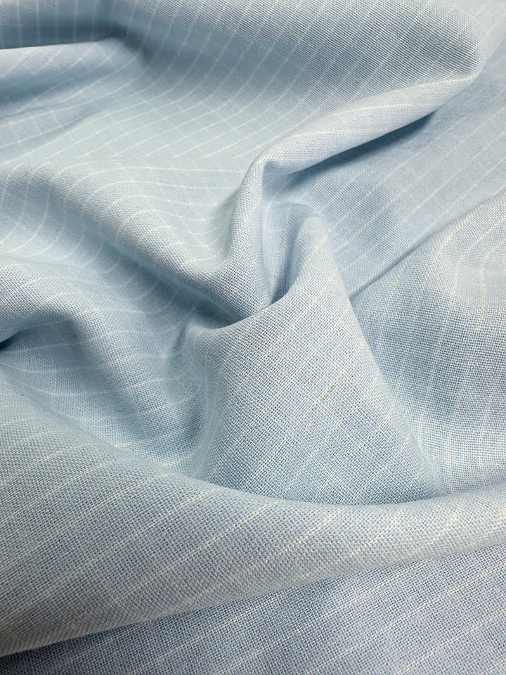 A close-up view of light blue fabric with a subtle pinstripe pattern. The high-quality Linen Rayon Grid - Baby Blue - 145cm by Super Cheap Fabrics appears soft and is slightly crumpled, showcasing its texture and smoothness, making it perfect for both home decor items and garment creations.