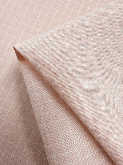 A close-up of Linen Rayon Grid - Pale Peach - 145cm fabric from Super Cheap Fabrics with a subtle white grid pattern. The fabric is slightly folded, showcasing its texture and design, perfect for garment creations or home decor.