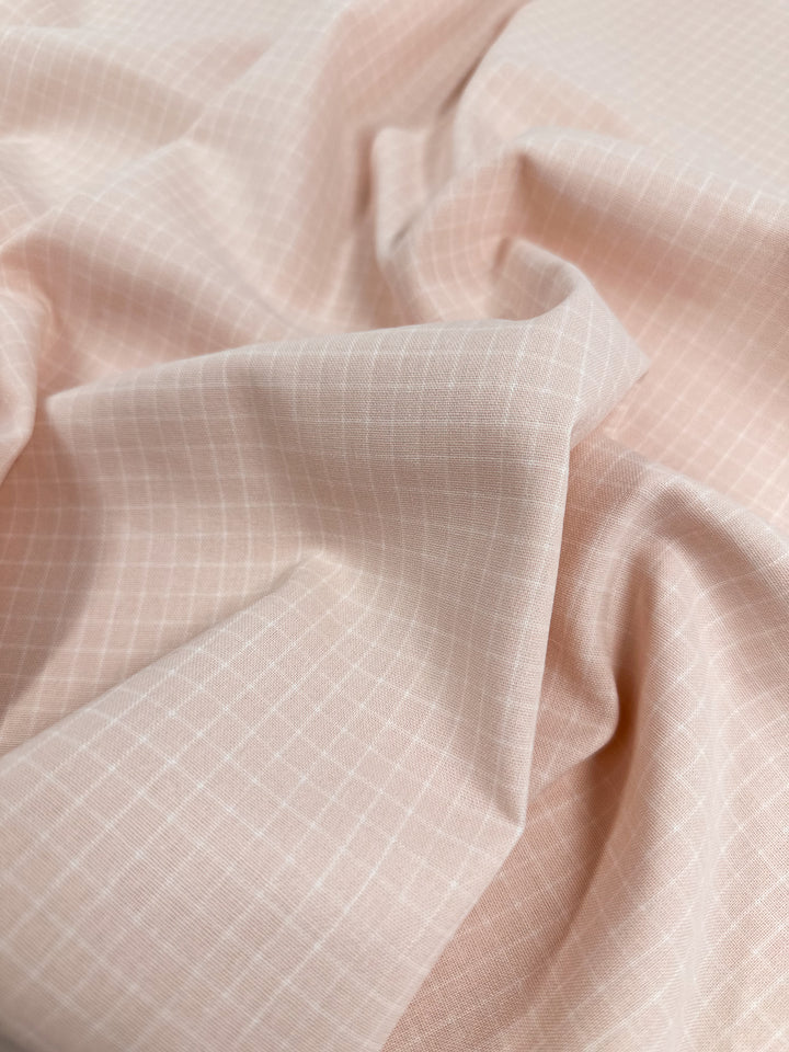 Close-up of a softly wrinkled pink printed Linen Rayon Grid - Pale Peach - 145cm fabric by Super Cheap Fabrics with a subtle white grid pattern. The material appears smooth and lightweight, ideal for garment creations or home decor, with gentle folds creating shadows and highlights that enhance the texture and design.