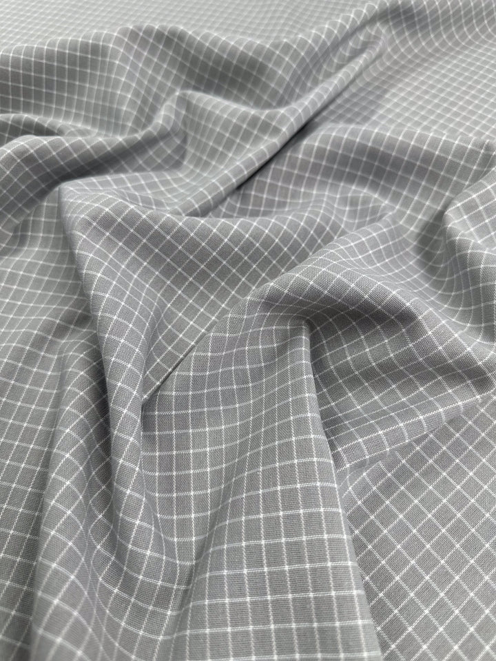 A close-up of a crumpled light gray fabric with a small white checkered pattern. The intricate grid design is uniform across the Linen Rayon Grid - Grey - 145cm by Super Cheap Fabrics, creating a textured and detailed appearance. This versatile material forms soft folds and shadows, perfect for home decor items.