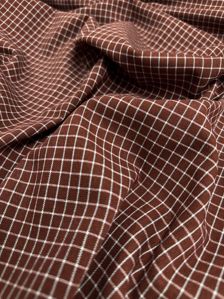 Close-up of a brown fabric with a white grid pattern. The fabric is slightly crumpled and draped, displaying the texture and design ideal for home decor fabric creations. This is the Linen Rayon Grid - Rosewood - 145cm from Super Cheap Fabrics.
